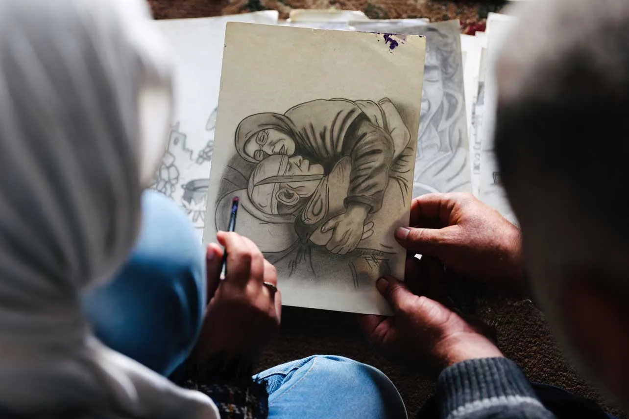 Un mujer muestra a otra el dibujo de una madre abazando el cuerpo sin vida de un hombre.